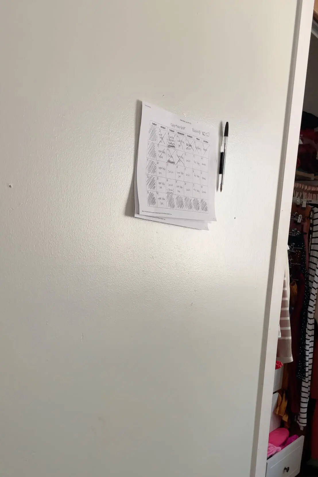 Close-up of a printed monthly workout calendar taped to a white wall, with a pen attached beside it. The calendar shows various workout routines with days marked off, illustrating a commitment to staying on track with fitness goals.